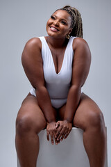 Excited plus size black woman in white bodysuit laughing, sitting in studio over white background,...