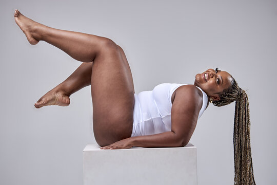 Plus size sexy happy feminine model in white beautiful bodysuit posing on cube, fat woman isolated on white studio background enjoy having overweight body, body positive and beauty concept.