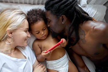 cute black daughter child have fun with diverse parents watching video on smartphone, dark skinned male and blonde lady relaxing on bed at home in cozy bedroom at day time or morning. top view