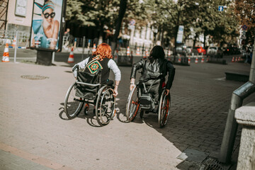Fototapeta premium Two girls in wheelchairs near an advertising banner with a smiling girl. Women with special need walk around Kiev near Khreshchatyk and admire the sights. Happy girl on a wheelchair with her friend