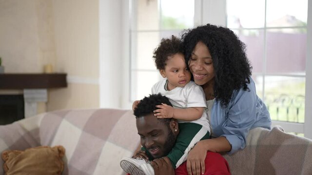 Happy African American mother hugging little son on shoulders of smiling father. Positive cheerful young millennial family enjoying weekend with boy at home in living room. Slow motion