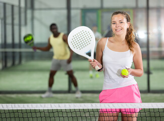 Portrait of young smiling sporty female in white t-shirt with padel racket and ball at tennis court