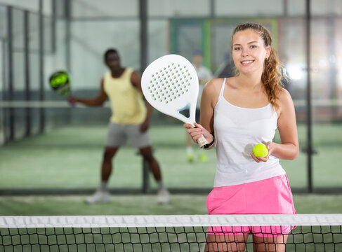 Portrait of young smiling sporty female in white t-shirt with padel racket and ball at tennis court