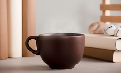 brown cup of coffee with hot steam, on the desk with shells and books, grey background