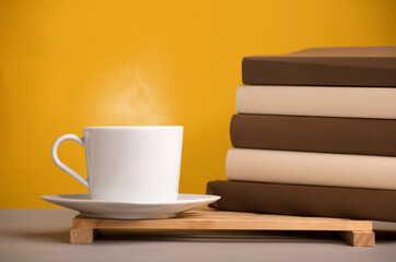 white cup of coffee with hot steam, on the desk with brown books, yellowish orange background