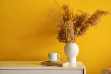 Vase with dry reeds, cup and book on shelf near yellow wall