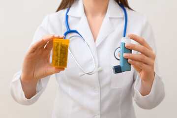 Female doctor with asthma inhaler and pills on light background