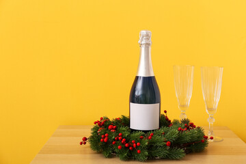 Bottle of champagne, glasses and Christmas wreath on table near color wall