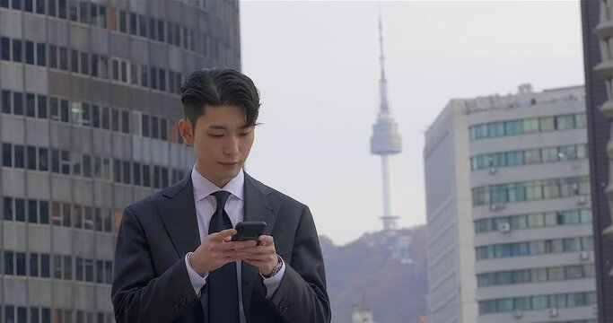 Well groomed korean male office employee in business attire typing messages on his mobile phone with high rise city buildings and YTN Namsam Tower in the background.