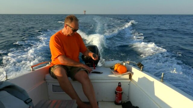 Caucasian man with sunglasses orange t-shirt and shorts on board of navigating motorboat holds steering tiller for direction while using cellphone. Slow-motion
