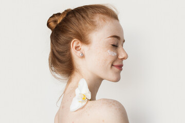 Side view of a smiling half naked woman applying face cream having an orchid on the soulder isolated over white background. Redhead caucasian woman. Skin care