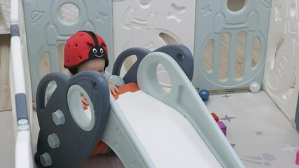 Cute toddler in baby safety helmet enjoy playing time in infants playpen with slide, 8 month old baby boy playing in safe activity center. High quality 4k footage