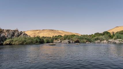 The Nile River flows between the banks with lush green vegetation, picturesque boulders. A sand dune against an azure sky. Ripples and reflections on blue water. Egypt