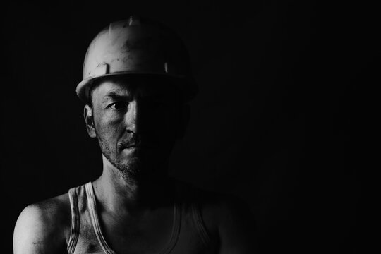 Dirty coal miner in a yellow hard hat on a dark background in a black and white photo.