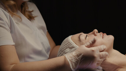 Obraz na płótnie Canvas Woman receiving anti-ageing facial massage in spa salon relax. Wellness body skin care face beauty treatment. Black background