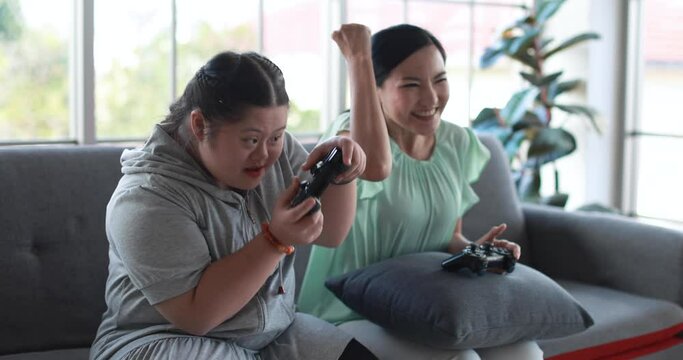 Cheerful daughter with down syndrome and Asian mother with joysticks playing videogame and celebrating win while having fun at home and sitting on sofa in living room