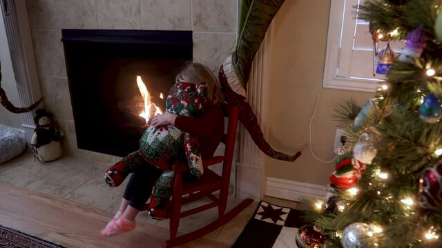Three-year old toddler girl sits by the fireplace and Christmas tree with her teddy bear