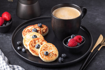 Cottage cheese pancakes with fresh with fresh raspberries and blueberries on dark ceramic plate. Cup of coffee and coffee maker