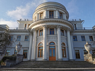 The main entrance to the Elaginoostrovsky Palace with a staircase and stone vases, trees with fallen leaves on the edges against a blue sky with clouds.