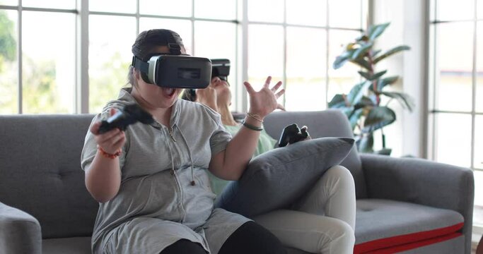 Girl with down syndrome and mother wearing VR goggles playing videogame with joysticks and celebrating victory while entertaining at weekend and sitting on sofa in living room
