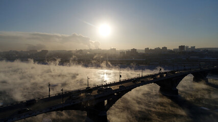 The old bridge over the Angara river. Steam is emitted from the water. Aerial drone flight. In winter, the Russian city is covered with ice and snow.