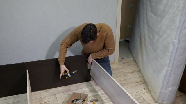 Young man in brown sweater assembles the bed with a screwdriver.