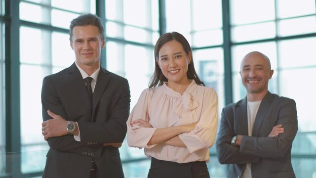Group of three business people are standing with crossed arms on window in office at high cityscape. They are giving cordinal smile.  