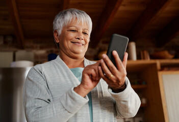 Happy multi-cultural elderly female typing a message on smartphone while standing in modern kitchen holding morning coffee.