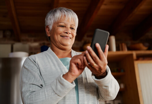 Happy multi-cultural elderly female typing a message on smartphone while standing in modern kitchen holding morning coffee.