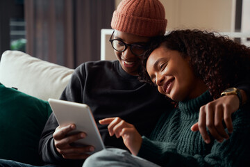 Cute adult couple, multi-ethnic, smiling at tablet on sofa in modern apartment