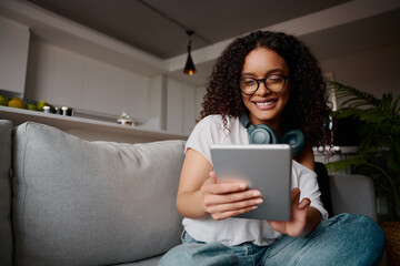 Multi-cultural young adult female using tablet on the sofa. Smiling with glasses on