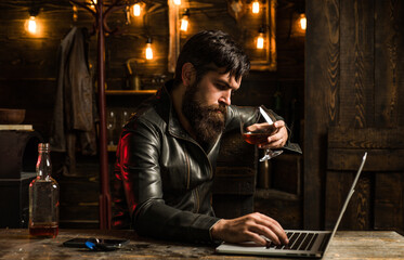 Businessman stressed with wiskey. Handsome man with a beard is sitting in a cafe with glass of whiskey and a laptop.