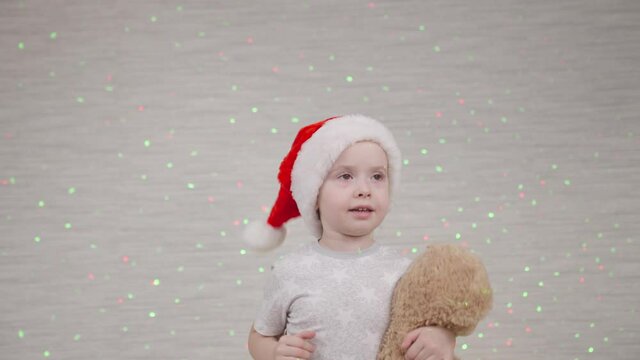 a little child is jumping on the sofa with a Santa hat on his head, the kid is playing with teddy bear toy in light of bright colorful lanterns, bokeh garlands on wall of room, happy childhood dream