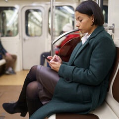 Tired african american woman office worker sitting on seat in subway train and using smartphone, making call or sending message, afro female employee getting home after work by public transportation