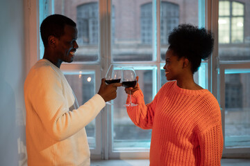 Young happy african couple enjoy romantic date at home, drink red wine, say love toast and clink wineglasses while standing by window, biracial man and woman celebrate anniversary of relationship