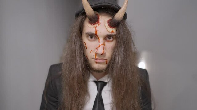 long haired white man with plastic horns on forehead and fake blood on face staring seriously