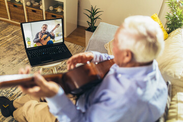 Senior man playing guitar at home using laptop for online lessons