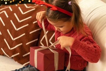 Little caucasian girl unpacks gift boxes at home by the Christmas tree.The girl is wearing a red...