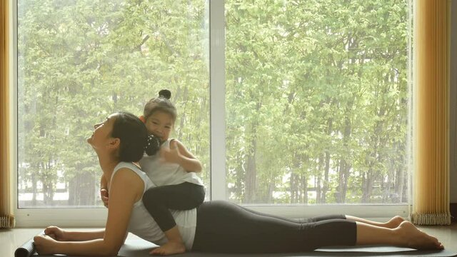 4K. Asian mom practice yoga exercise at home with a adorable daughter sitting on mother back. training during quarantine self-isolation. leisure healthy lifestyle. healthy family's activity in the mor