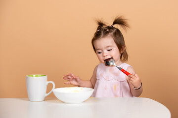 Baby girl with spoon on studio, isolated on beige yellow. Cute funny babies eating, baby food,