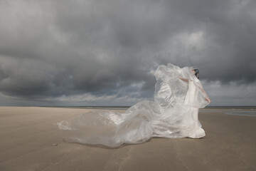 Fine Art portrait of  Woman in white dress on the beach wrapped in plastic sheet in storm and sunlight
