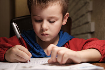 Child Is Doing Homework In The Light Of The Lamp In The Evening. A Boy Sits at a Table and Writes.