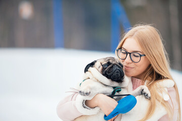 Satisfied happy pug dog in arms of young woman owner in winter in park for walk