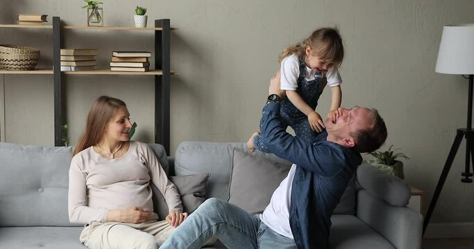 Smiling mother expecting baby sit on couch watch father playing with daughter toddler act flying airplane lift happy kid in air. Family of 3 pregnant mom dad preschool girl spend leisure time together