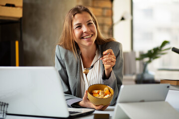 Businesswoman in office having healthy snack. Young woman eating fruit while having a video call.