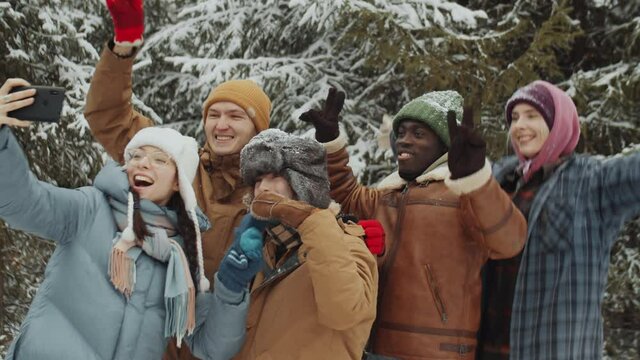 Company of young happy multiethnic friends smiling and posing together for smartphone camera while taking selfie in winter forest