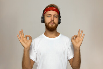 Attractive manful Caucasian guy with blond beard wearing wireless headphones and red bandana,...