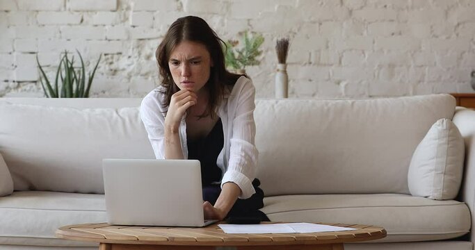 Distressed woman sit on couch look at laptop screen, pay calculates bills feels shocked by debt, summarizing utilities experiences financial problem, lack of money. Crisis, overspend, high fee concept