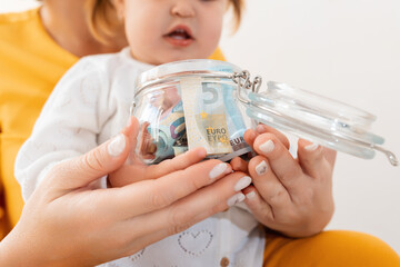 A mother and her daughter together hold a glass jar filled with money. Close up of hands. The concept of finance and accumulation of funds