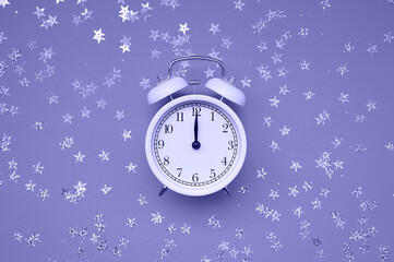 White alarm clock on paper background with silver confetti stars, glitter. The trending color of 2022. Veri peri